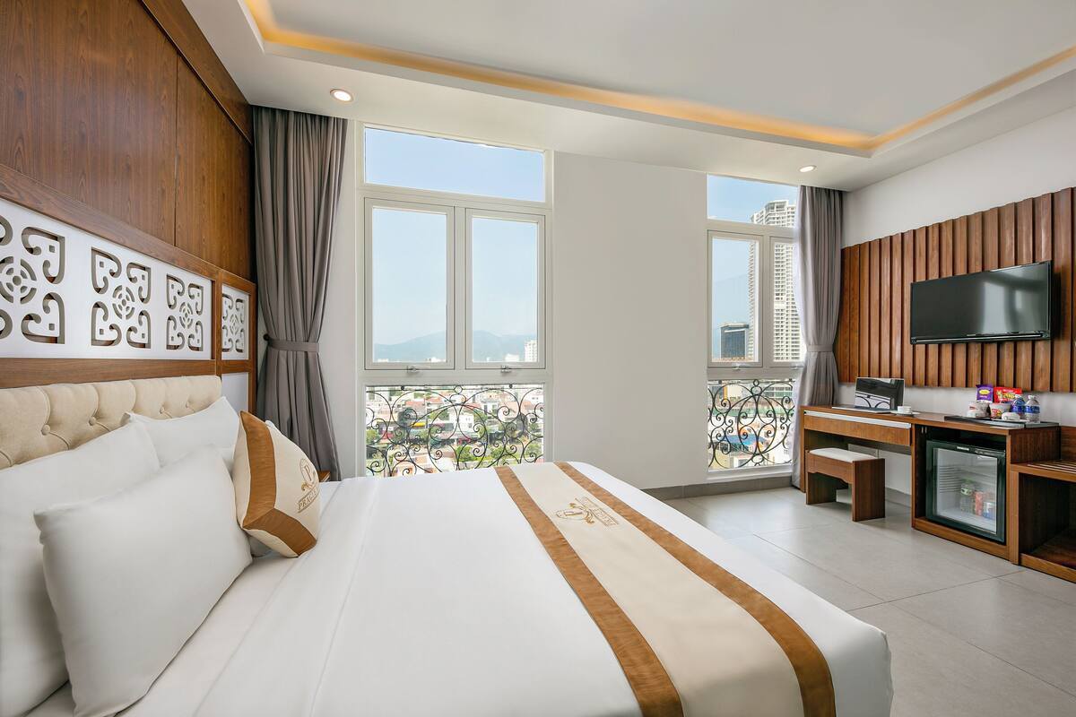 A hotel room with a large bed, a desk with a television, and a balcony with a city view.