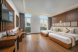 Ein Hotelzimmer mit Holzboden, einem Schreibtisch mit Stuhl, zwei Betten, einem an der Wand montierten Fernseher und großen Fenstern mit Blick auf umliegende Gebäude.