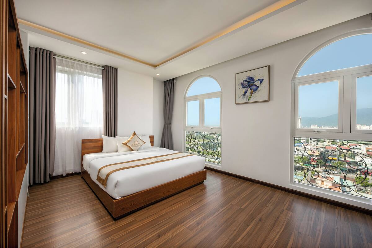 A hotel room with a large bed, wooden flooring, a balcony with a city view, and a painting on the wall.