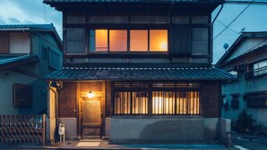 A traditional Japanese house with a tiled roof, a wooden entrance gate, and a lit interior.