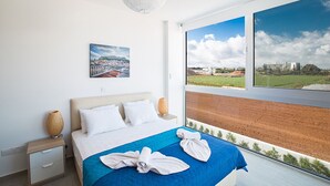 Quarto de hotel moderno com uma cama grande, mesa de cabeceira, luminária e pintura de um horizonte urbano.