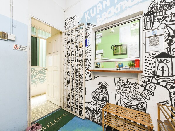 Una habitación con felpudo de bienvenida, un banco, una pared con arte blanco y negro, una ventana con vistas y una puerta con letrero que dice 'no se permiten zapatos'.