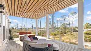 Una terraza de madera con un sofá de mimbre y cojines, con vistas a un paisaje con árboles y un edificio distante.