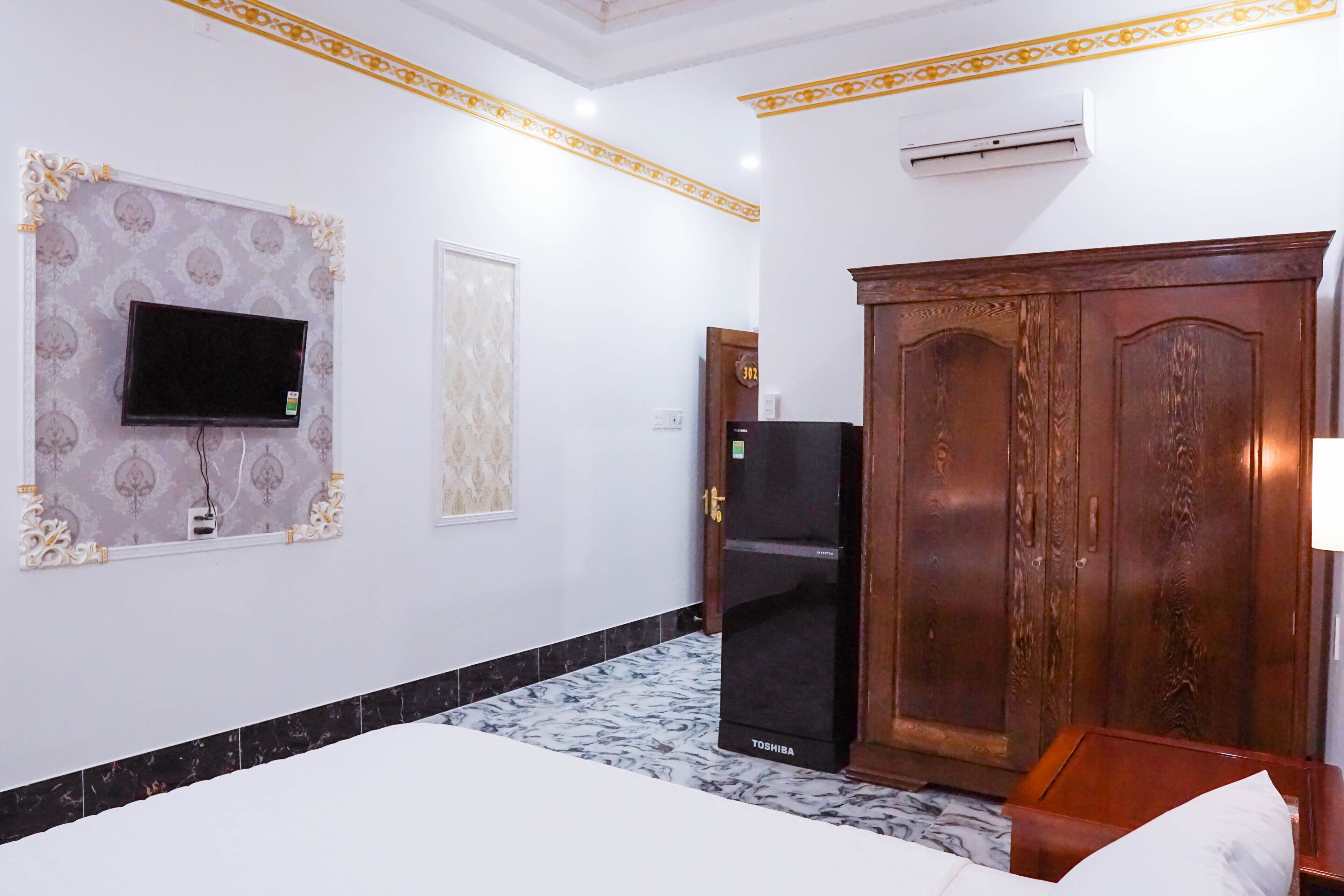 A hotel room with a bed, a television mounted on the wall, a refrigerator, and a wooden wardrobe.