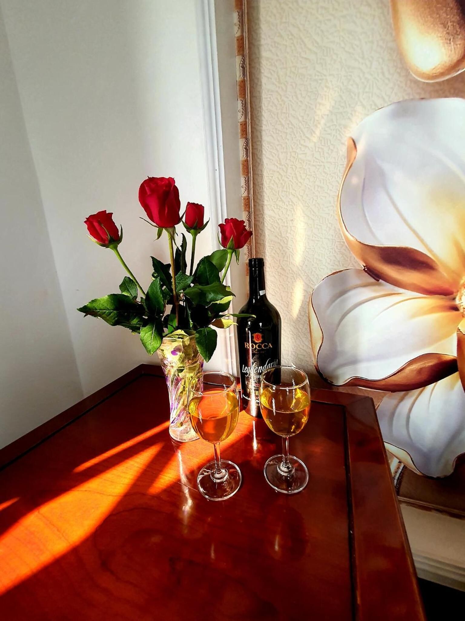 A table with a vase of red roses, a bottle of wine, and two wine glasses.