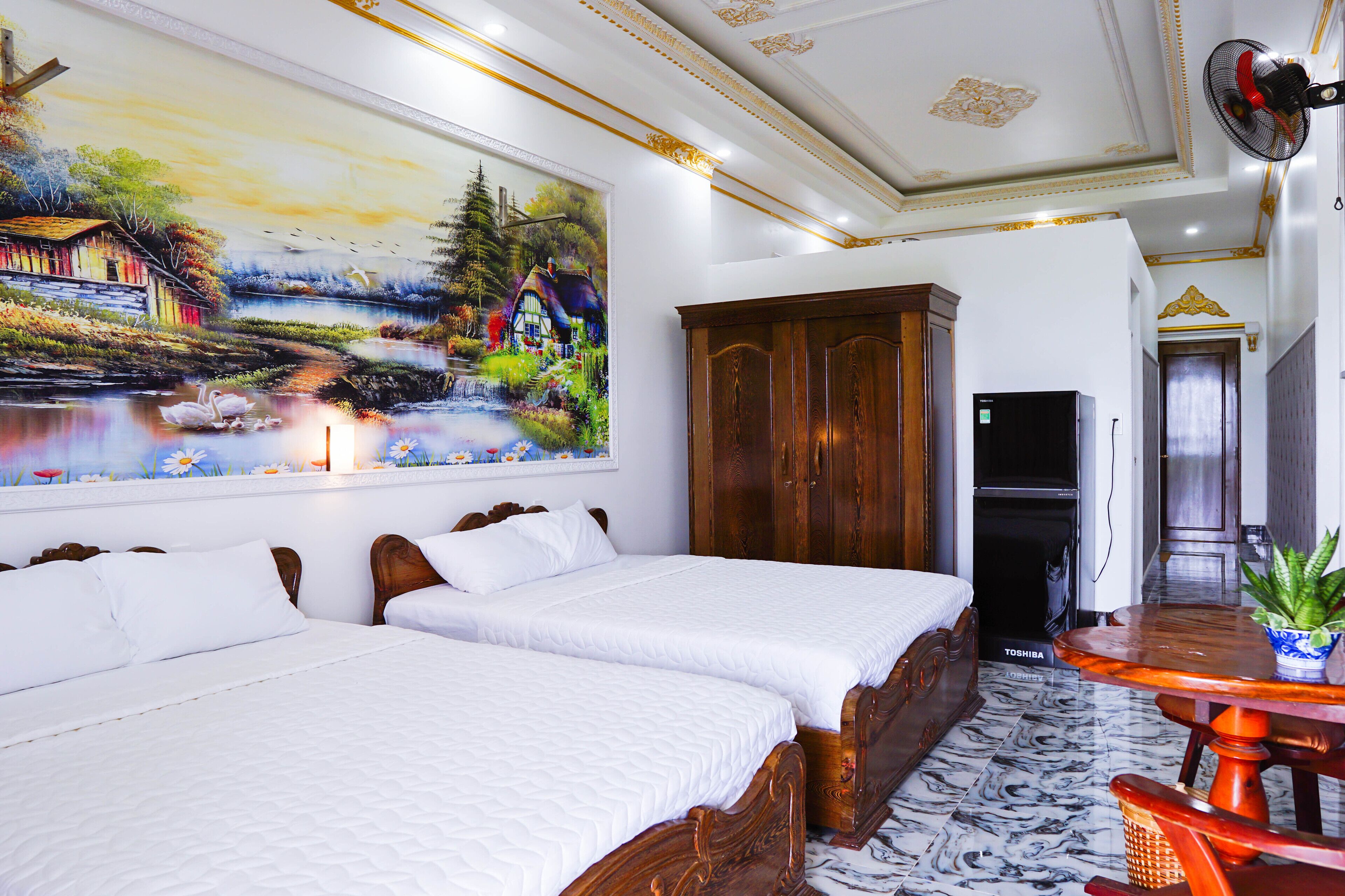 A hotel room with two beds, a wooden wardrobe, a dining table with a plant, and a mural on the wall.