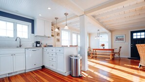 A spacious kitchen with white cabinetry, a dishwasher, and a trash can. There is a dining area with a wooden table and chairs.