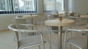 Un comedor con mesas redondas y sillas de metal, ventanas amplias con persianas y suelo de baldosas.