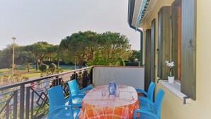 Un balcón exterior con una mesa y sillas, una planta en maceta y vistas a árboles y un jardín.