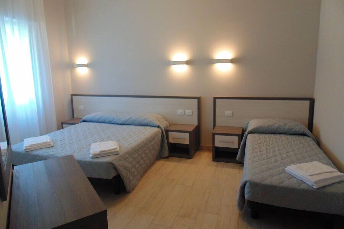 A hotel room with two single beds, each with a bedside table and lamp. The room has a wooden floor and light-colored walls.