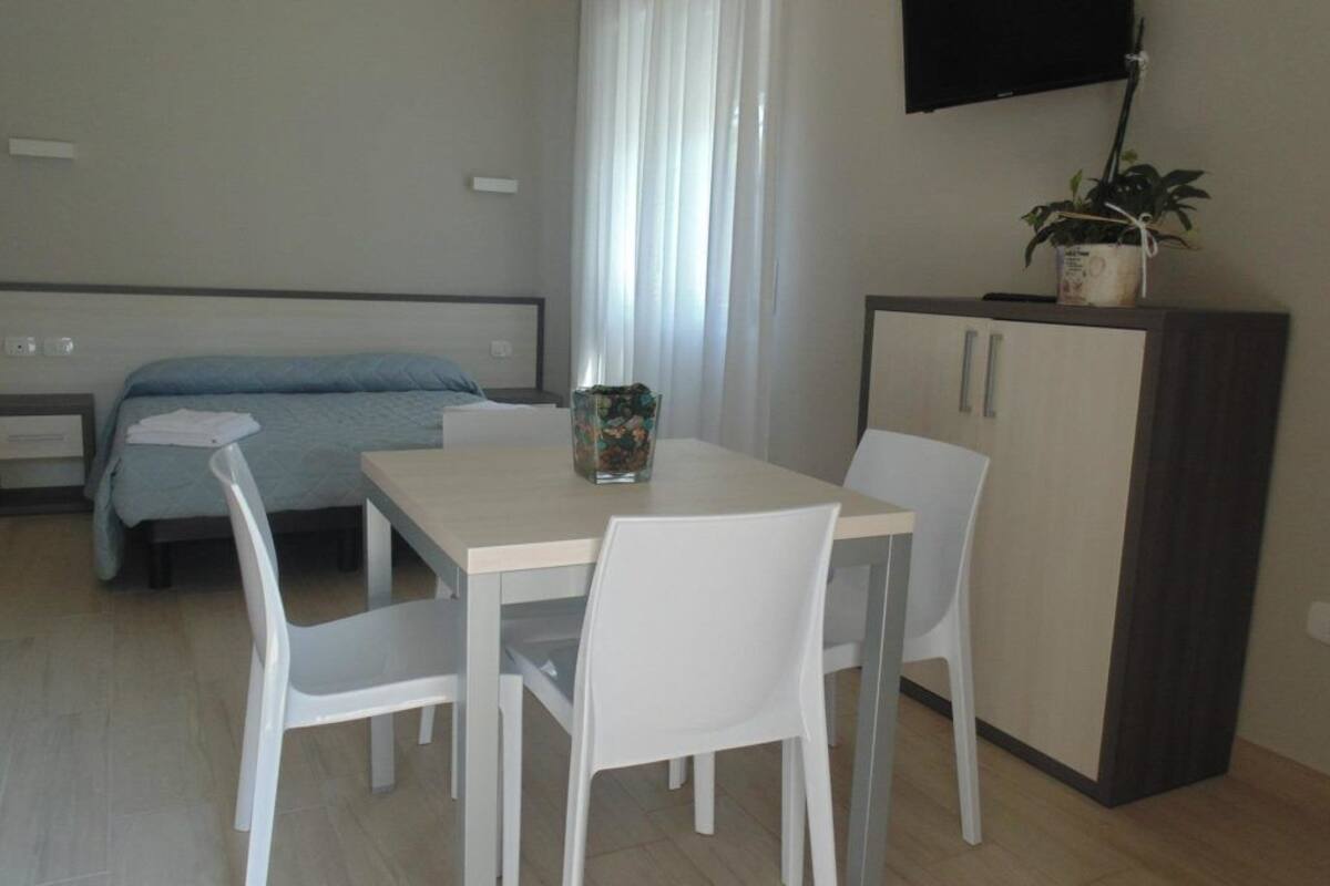 A room with a bed, a table with chairs, a TV mounted on the wall, and a cabinet with a plant on top.