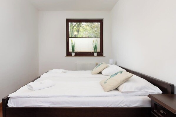 Una cama bien tendida con sábanas blancas y dos almohadas, una ventana con persianas y un radiador debajo.
