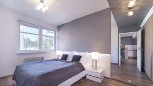 Ein modernes Schlafzimmer mit einem großen Bett, Nachttischlampen und Blick auf die Küche durch eine Glastür.