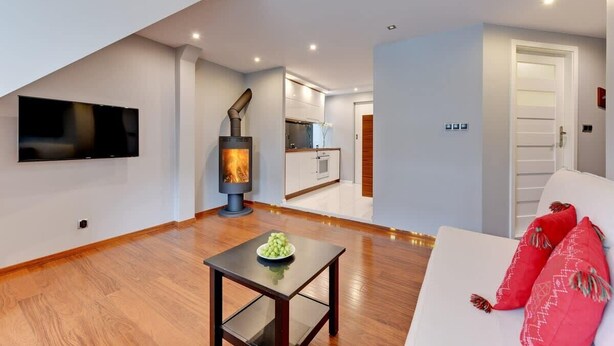 Una sala de estar moderna con televisor de pantalla plana, chimenea de leña, una mesa de comedor con un cuenco de fruta y un sofá blanco con cojines rojos.
