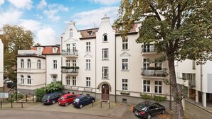 Una hilera de edificios residenciales blancos de varios pisos, con balcones, coches aparcados y un árbol en primer plano.