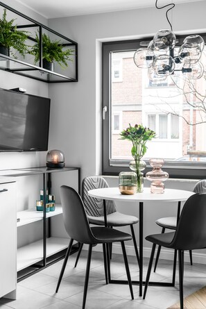 Un comedor moderno con una mesa blanca, sillas negras, una lámpara de cristal y un amplio ventanal con vistas a edificios.