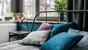 A bed with multiple pillows, a bedside table with a lamp, and a window with a view of buildings and a plant. - Green and blue apartments (Gdansk)