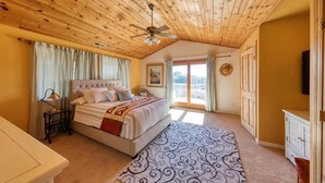 Ein Schlafzimmer mit Holzboden, einem Bett mit Kopfteil, einem Nachttisch, einem Fenster mit Vorhängen, einer Tür und Blick ins Freie.