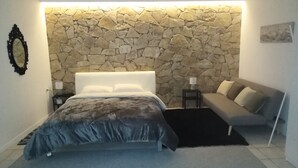 A bedroom with a stone wall, a bed with a grey duvet, a grey sofa, a small table with a lamp, and a mirror on the wall. - Stone Lobby 11 (Markopoulo Mesogaias)