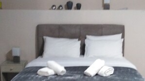 A neatly made bed with white linens and towels, a bedside table with a lamp, and a wall-mounted shelf with a few items. - Stone Lobby 11 (Markopoulo Mesogaias)