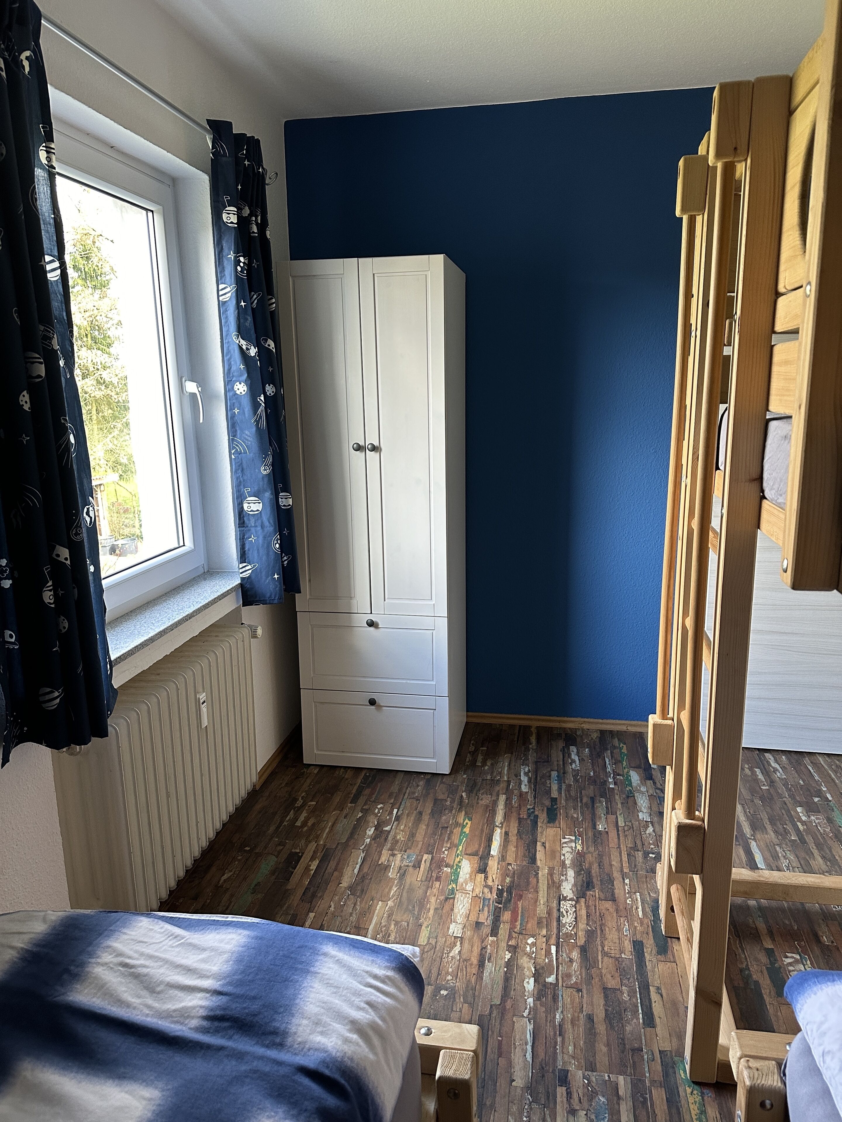 A room with a bunk bed, a white wardrobe, a radiator, and a window with curtains.