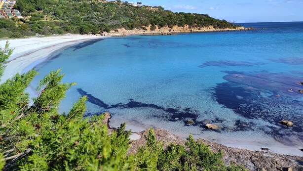 Un paisaje costero con aguas azules cristalinas, una playa arenosa y una colina con edificios.