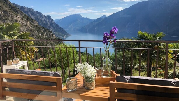 Ein Balkon mit Holzmöbeln, einer Blumenvase und Blick auf einen See, der von Bergen umgeben ist.