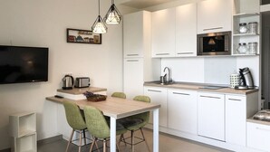 Eine moderne Küche mit weißen Schränken, einem eingebauten Backofen und einer Kaffeemaschine. Ein Esstisch mit Stühlen und ein an der Wand befestigter Fernseher sind ebenfalls vorhanden.