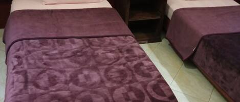 Habitación de hotel con dos camas, cada una con cubrecama y almohadas moradas a juego. Entre las camas hay una mesita de noche de madera con un control remoto y un vaso encima. El piso es de baldosas y la pared es roja con un detalle dorado.