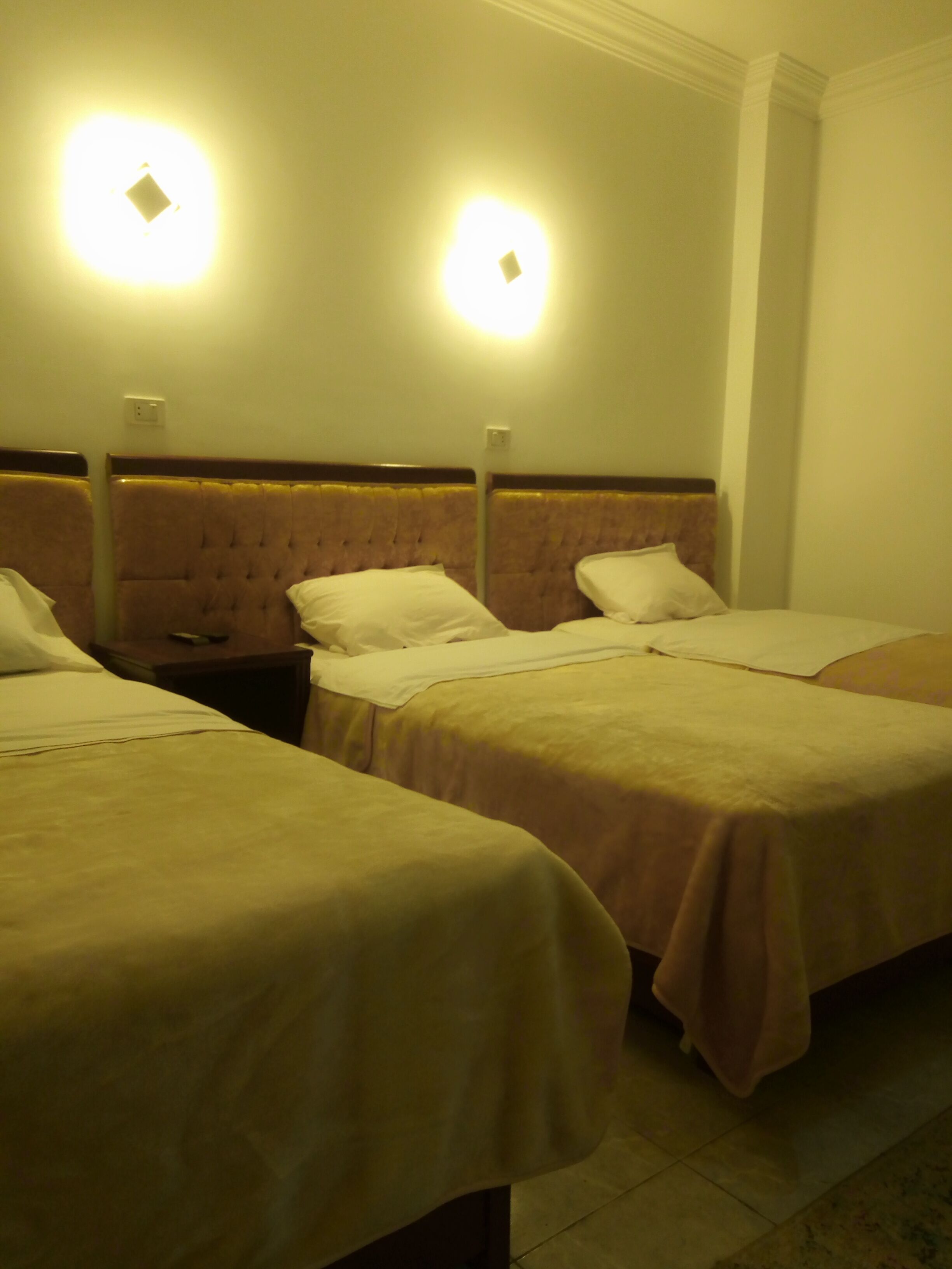 A hotel room with three beds, each with a headboard and a pillow. There are wall-mounted lights above the beds.