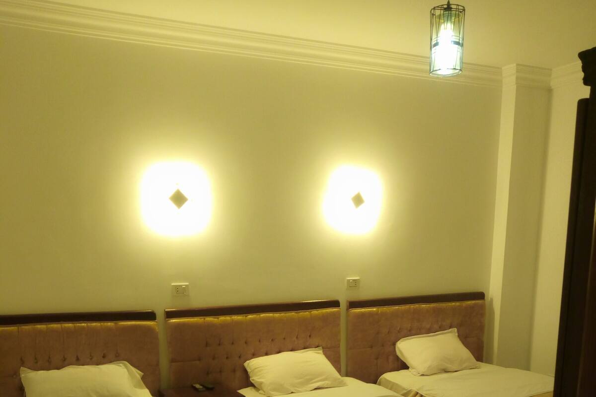 A hotel room with three single beds, each with a headboard and a bedside table. There are two wall-mounted lights above the beds.