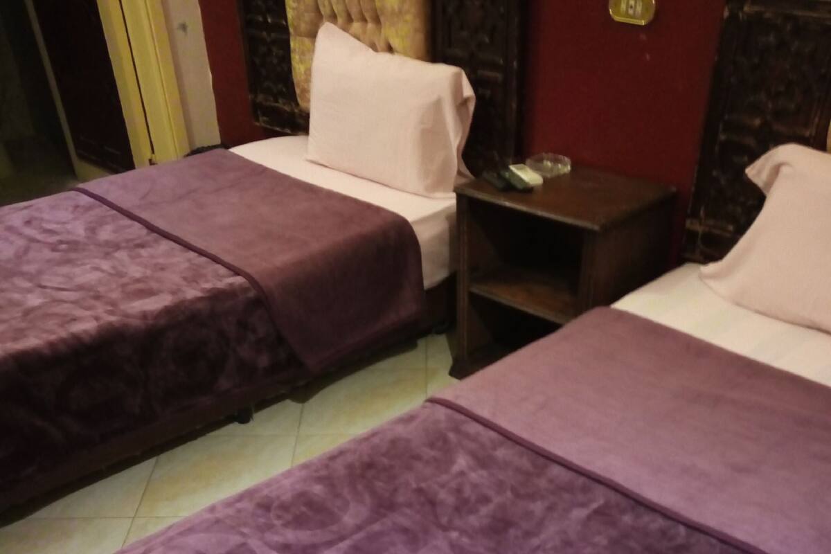 A hotel room with two beds, each with a purple bedspread and a white pillow. There is a wooden nightstand with a lamp and a small table beside the beds.