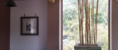 A dining area with a table set for two, a hanging light fixture, a window with a view of greenery, and a wall-mounted clock.