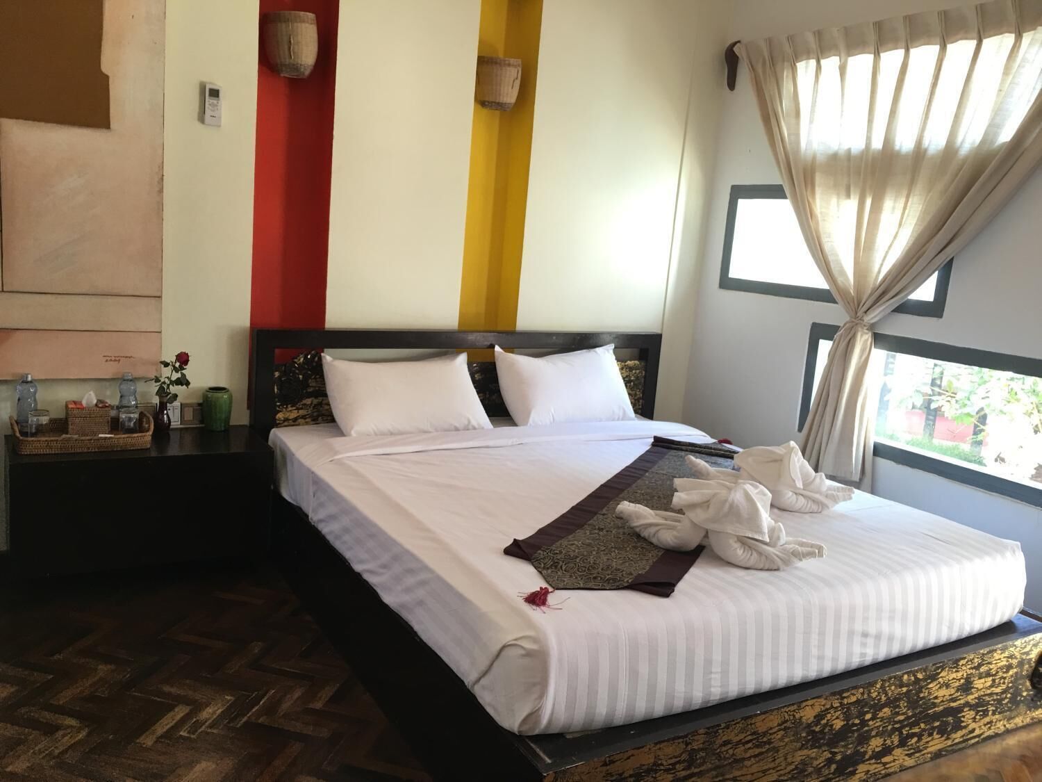 A hotel room with a large bed, a wooden headboard, a bedside table with a vase and a tray, and a window with curtains.