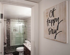 A bathroom with a glass shower enclosure, a toilet, and a vanity with a marble countertop. There is a wall-mounted sign that reads "Off Happy Day". - The BEST Old Town Scottsdale location!!1 bd /1bth beautiful mid century condo. (Scottsdale)