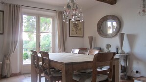 Comedor con mesa y sillas de madera, una araña, un ventanal con cortinas, un espejo y una estatua sobre un pedestal.