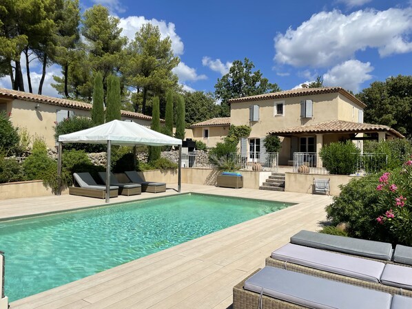 A pool area with lounge chairs and a canopy, surrounded by trees and a building with a tiled roof.