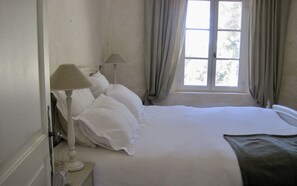 A neatly made bed with white linens, a bedside lamp, and a window with curtains.