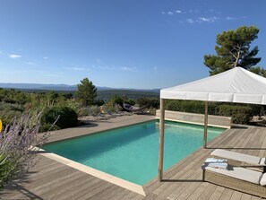 A wooden deck with a swimming pool, lounge chairs, a canopy, and a view of distant mountains.