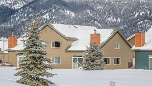 Una casa de dos pisos con tejado nevado y una chimenea, rodeada de árboles y montañas cubiertas de nieve.