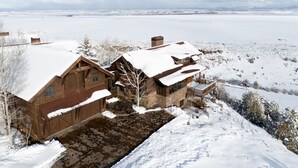 A wooden cabin with a snow-covered roof, situated in a snowy landscape with a clear view of distant mountains.