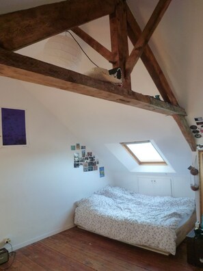Ein Schlafzimmer mit schrägen Wänden, Holzbalken, einem Oberlicht, einem Bett mit floralem Überwurf und einer Wand mit Fotos.