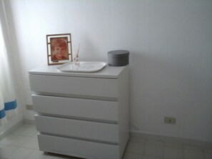 Uma cômoda branca com gavetas e um pequeno recipiente cinza em cima, um quadro emoldurado e uma vela em um castiçal de vidro.