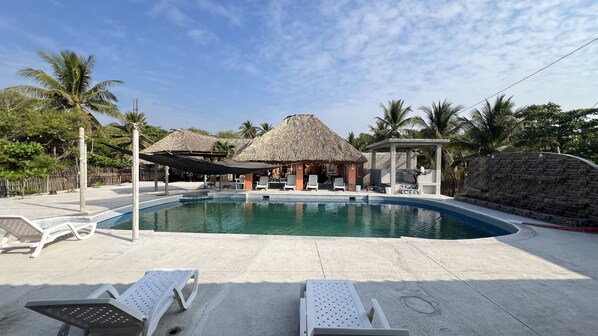 A pool area with lounge chairs, a thatched-roof structure, and palm trees.