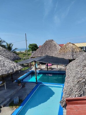 A pool area with thatched-roof structures, a clear sky, and a view of the ocean.