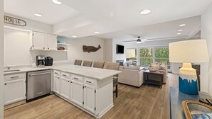 Una cocina moderna con armarios blancos, lavavajillas e isla central. Hay una zona de comedor con sofá y televisor.