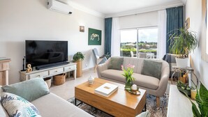 Sala de estar moderna com sofá, mesa de centro e televisão. Há uma varanda com vista para a paisagem.