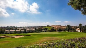 Um campo de golfe com céu claro, casas ao fundo e palmeiras.