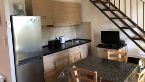 A compact kitchen with a refrigerator, stove, and microwave. There is a dining table with chairs and a television on a stand. A staircase is visible in the background. - Corsica: apartment in residence *** 6 people - swimming pool at 100m beach (Linguizzetta)