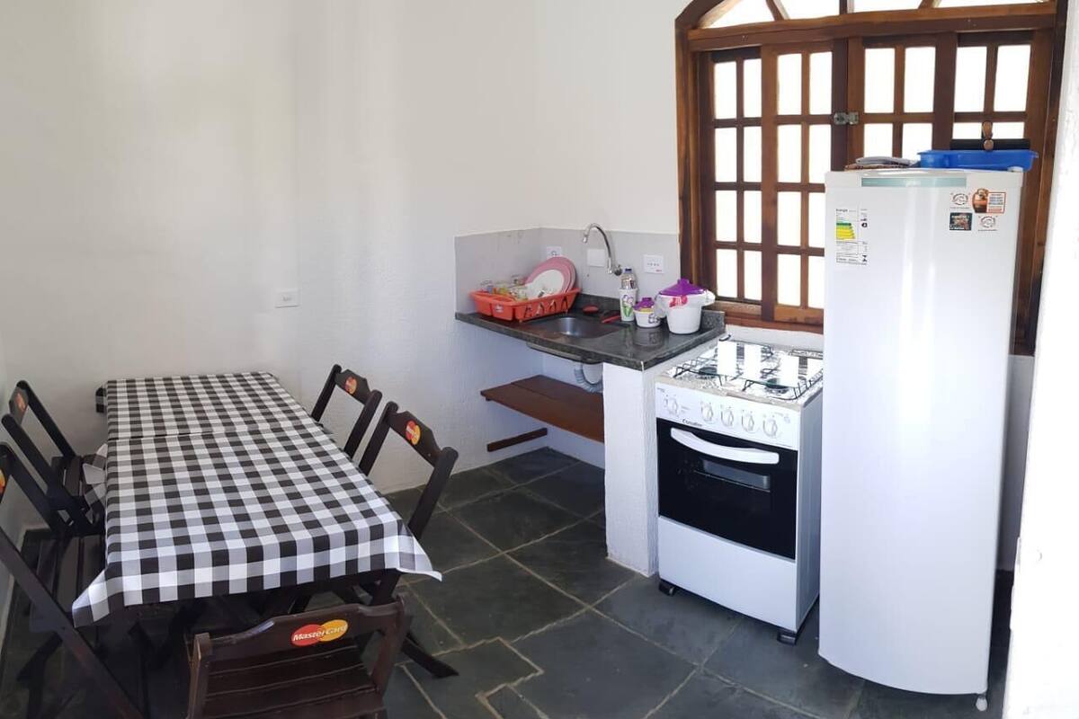 Eine kleine Küche mit Gasherd, Spüle und Kühlschrank. Ein karierter Tisch steht mit Stühlen bereit, und ein Fenster mit Holzjalousien sorgt für Licht.